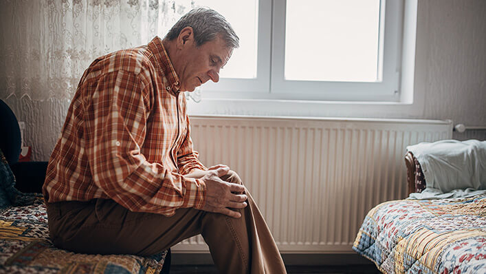 A man sits on the edge of a bed rubbing his sore knee.