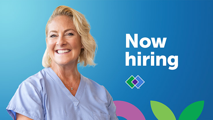 A woman wearing scrubs smiles in front of a background that says “now hiring.”