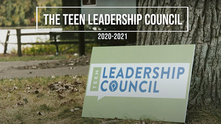 A picture of the Teen Leadership Council logo on a sign placed against the trunk of a tree. 