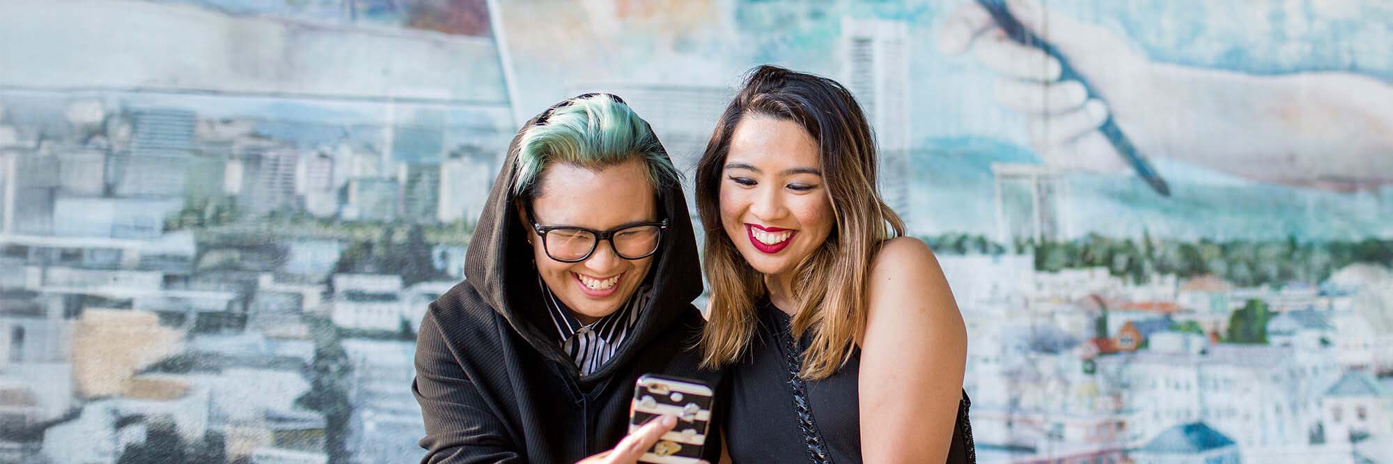 Two friends in their twenties laugh over a smartphone while sitting in front of a painted mural of a city scene.