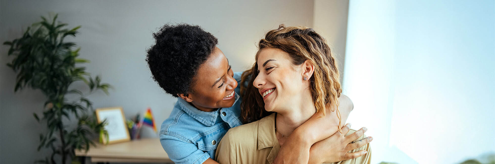 Two women gaze lovingly at each other in their home which has a rainbow decoration in the background.