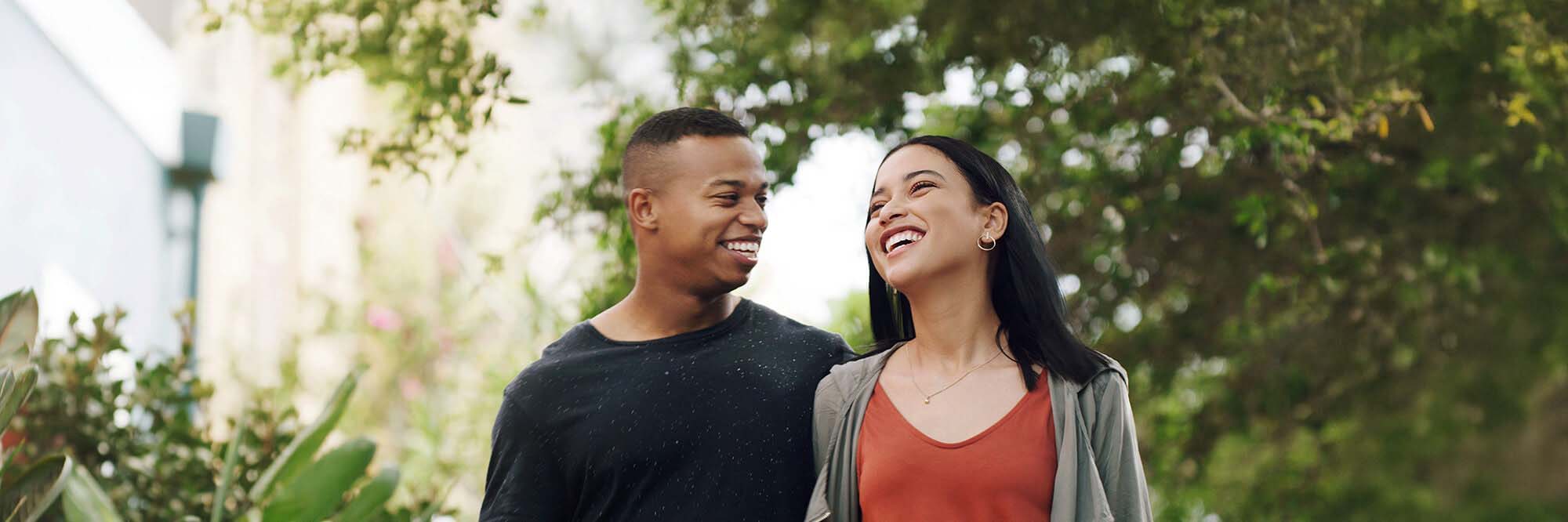 A young couple smile as they walk outside through trees and greenery.
